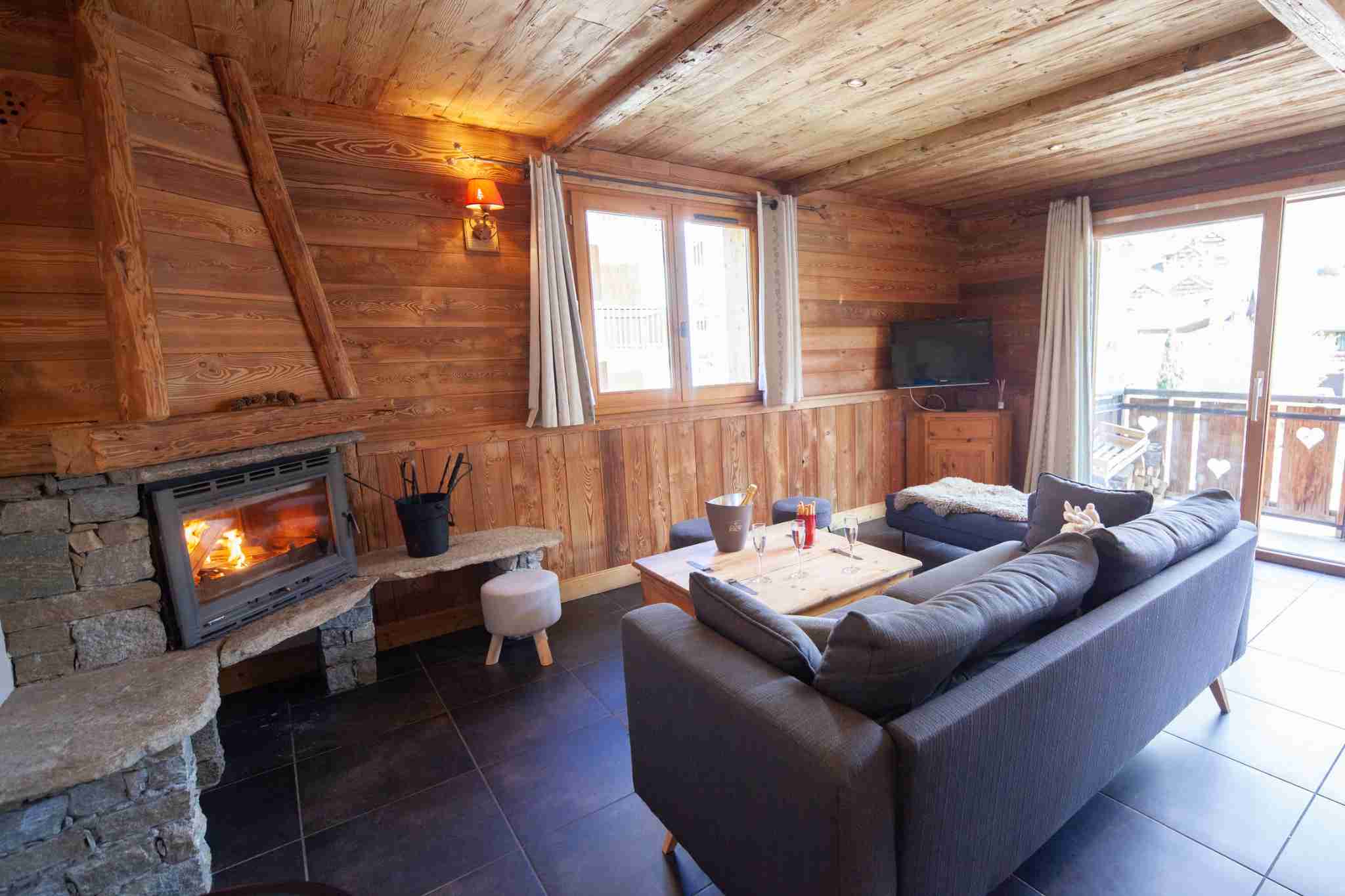 Salon du chalet egua à valloire, en bois avec cheminée, canapé gris et accès au balcon.